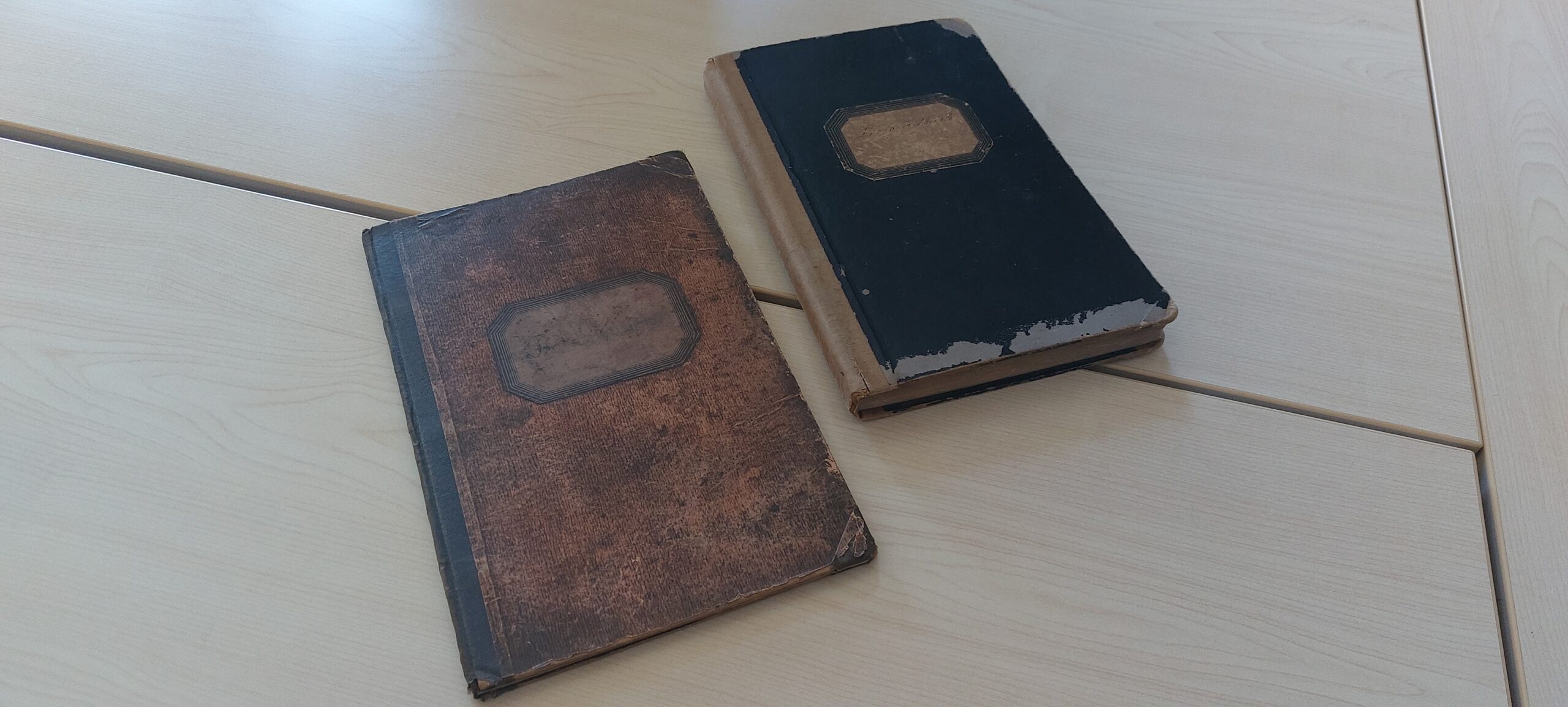 Zwei alte, abgenutzte Bücher liegen auf einem hellen Holztisch. Das linke Buch hat einen braunen Einband, das rechte einen schwarzen.