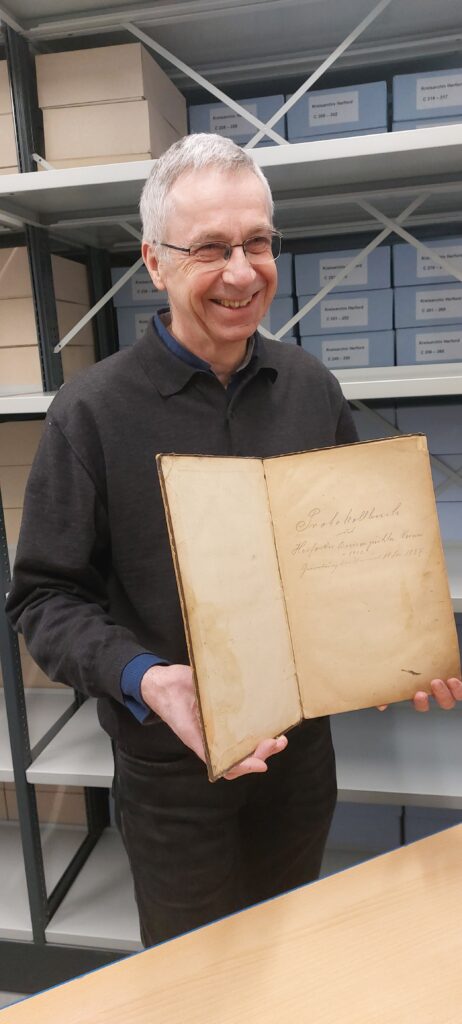 Udo Rolfsmeier im Archiv hält ein aufgeschlagenes, historisches Protokollbuch in die Kamera. Im Hintergrund sind Regale mit Archivkartons des Kreisarchivs Herford zu sehen.