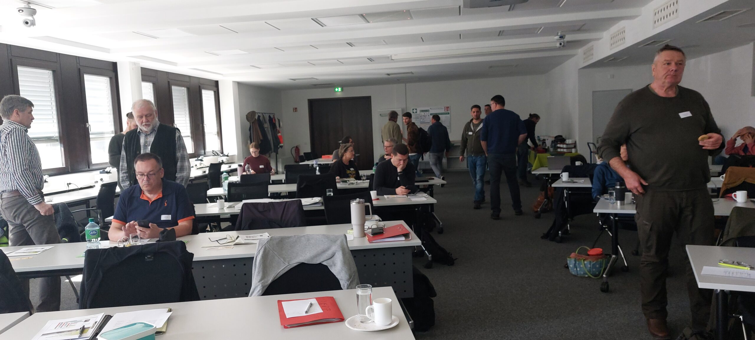 Ein Weitwinkel-Foto eines Konferenz- oder Schulungsraums, in dem sich mehrere Personen aufhalten, von denen einige an Tischen sitzen und andere stehen oder gehen, mit vielen Tischen, Stühlen und Papieren.