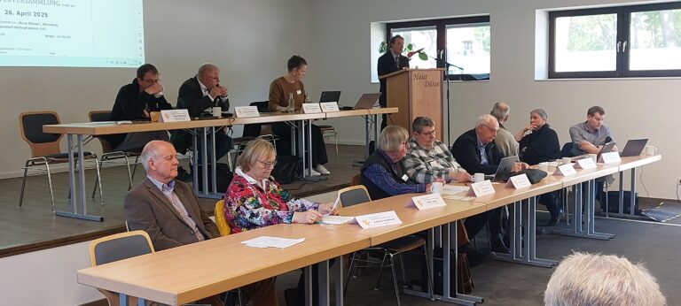 Ein Tagungssaal im Versuchs- und Bildungszentrum Landwirtschaft Haus Düsse während einer Versammlung am 26. April 2025. Auf einem Podium sitzen Vorstandsmitglieder an Tischen mit Namensschildern, während ein Redner an einem Holzpult mit der Aufschrift „Haus Düsse“ spricht. Im Vordergrund sitzen die Obleuteeiner an langen Tischreihe.