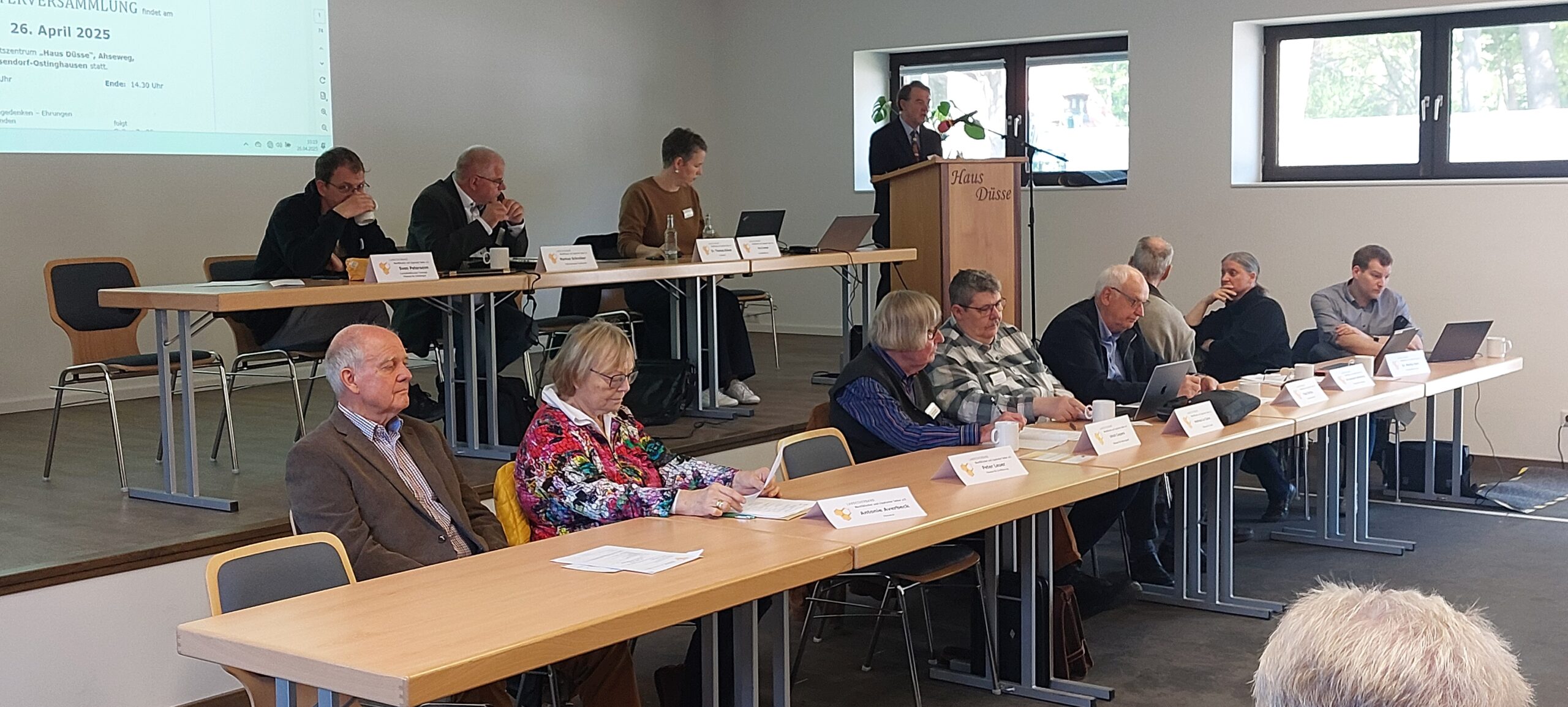 Ein Tagungssaal im Versuchs- und Bildungszentrum Landwirtschaft Haus Düsse während einer Versammlung am 26. April 2025. Auf einem Podium sitzen Vorstandsmitglieder an Tischen mit Namensschildern, während ein Redner an einem Holzpult mit der Aufschrift „Haus Düsse“ spricht. Im Vordergrund sitzen die Obleuteeiner an langen Tischreihe.