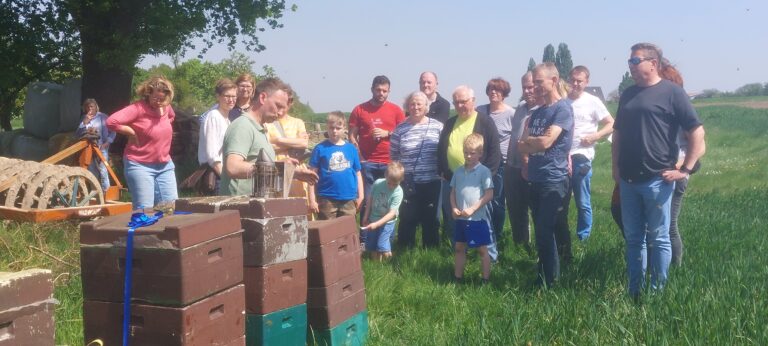 Ein Imker steht im Freien an mehreren aufgestapelten Bienenbeuten und erklärt einer Gruppe von interessierten Erwachsenen und Kindern die Arbeit mit den Bienen. Auf einer der Beuten steht ein rauchender Smoker.