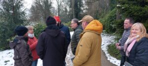 Eine Gruppe von sieben Erwachsenen steht im Winter auf einem leicht verschneiten Parkweg zusammen und unterhält sich.