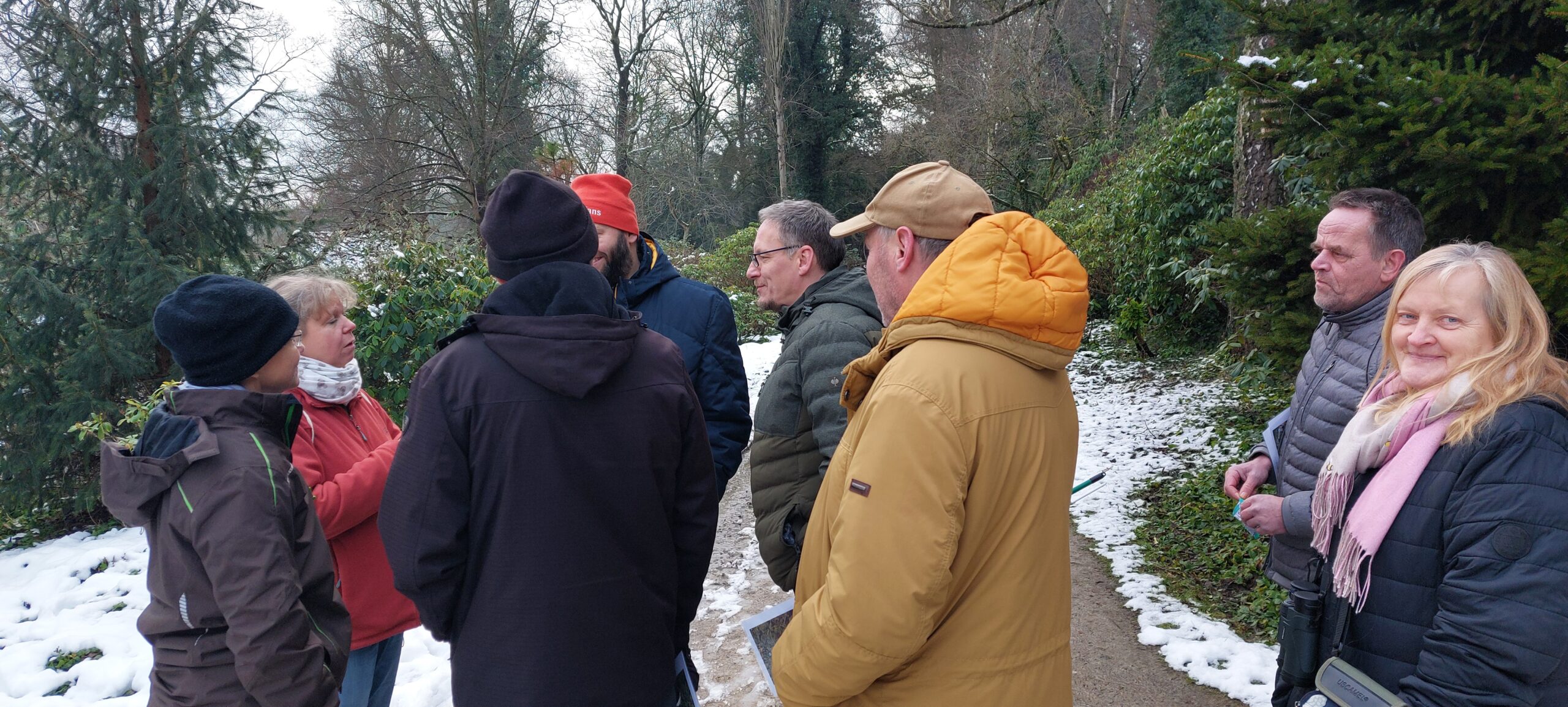 Eine Gruppe von sieben Erwachsenen steht im Winter auf einem leicht verschneiten Parkweg zusammen und unterhält sich.