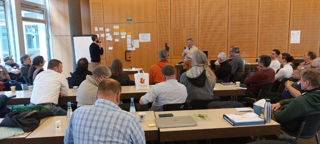 Ein Moderator präsentiert an einer mit Post-its beklebten Holzwand vor einer Gruppe von Teilnehmern in einem Seminarraum.