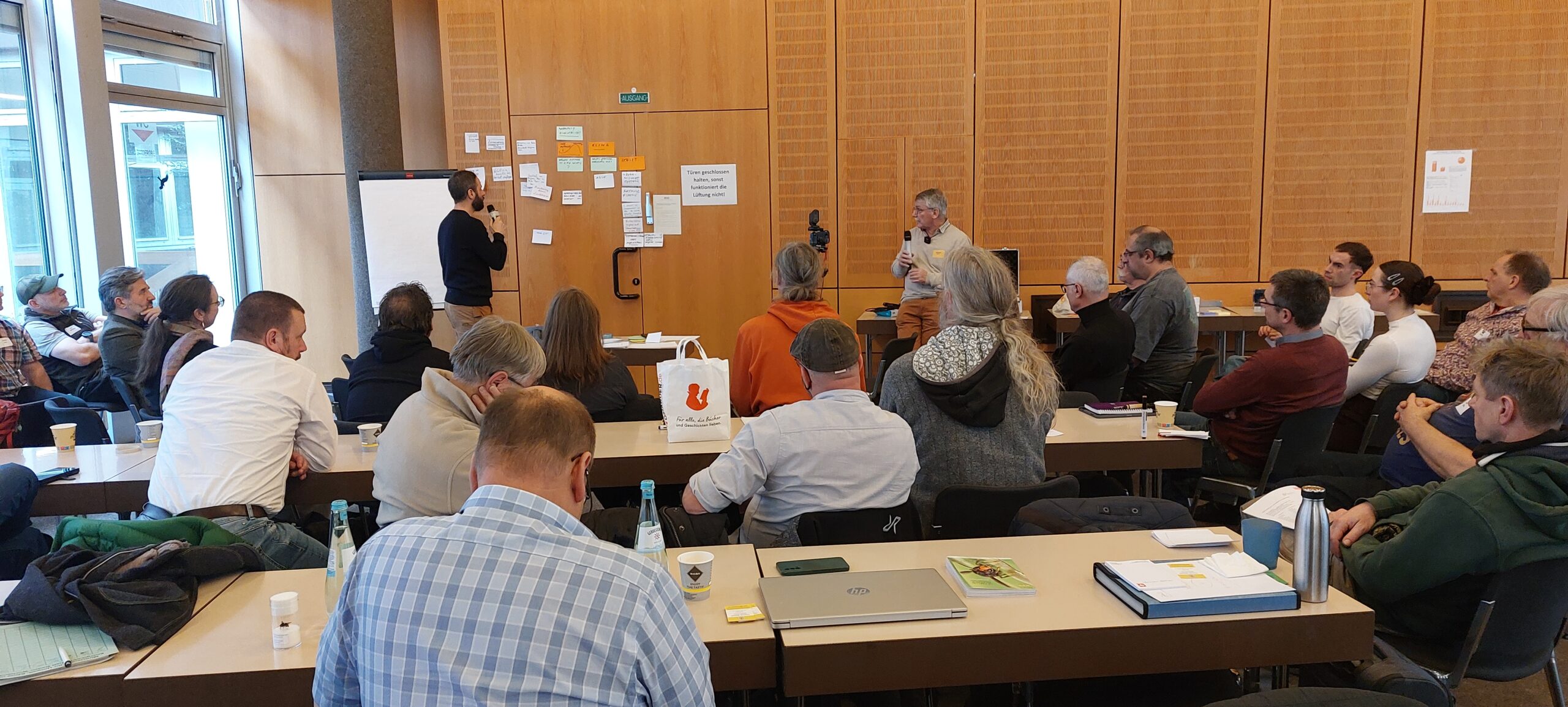Ein Moderator präsentiert an einer mit Post-its beklebten Holzwand vor einer Gruppe von Teilnehmern in einem Seminarraum.