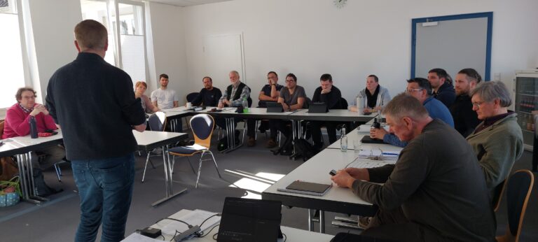 Eine Gruppe von Teilnehmern sitzt an einer U-förmigen Tischanordnung in einem hellen Seminarraum und hört einem Referenten zu, der im Vordergrund mit dem Rücken zur Kamera steht. Die Teilnehmer sind gemischten Alters und nutzen Tablets, Notizblöcke und Smartphones.