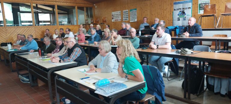 Ein vollbesetzter Schulungsraum mit Holzvertäfelung, in dem zahlreiche Teilnehmer in mehreren Reihen an Tischen sitzen und aufmerksam einem Vortrag folgen. An der Rückwand hängen Informationstafeln zu Bienen und Naturthemen.