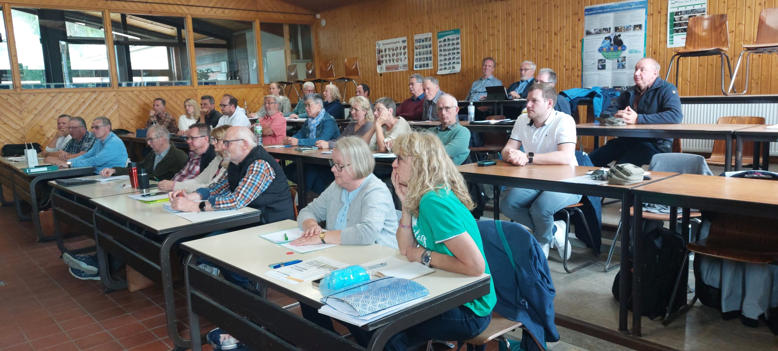 Ein vollbesetzter Schulungsraum mit Holzvertäfelung, in dem zahlreiche Teilnehmer in mehreren Reihen an Tischen sitzen und aufmerksam einem Vortrag folgen. An der Rückwand hängen Informationstafeln zu Bienen und Naturthemen.