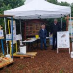 Eine Frau und ein Mann stehen lächelnd an einem Informationsstand im Freien unter einem weißen Pavillon. Links ist ein großes Insektenhotel zu sehen, davor ein Tisch mit Honiggläsern. Rechts steht ein Aufsteller zur Asiatischen Hornisse und eine Informationstafel zu Wildbienen.