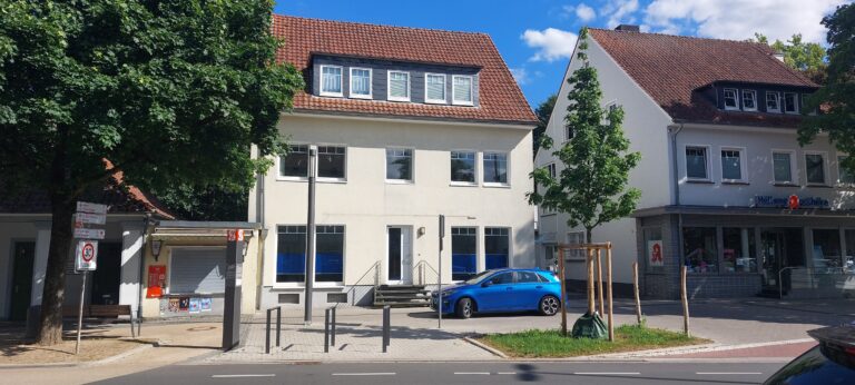 Helles zweistöckiges Gebäude mit Mansarddach neben einer Apotheke an einer Straße mit Parkplätzen und einem blauen Auto.