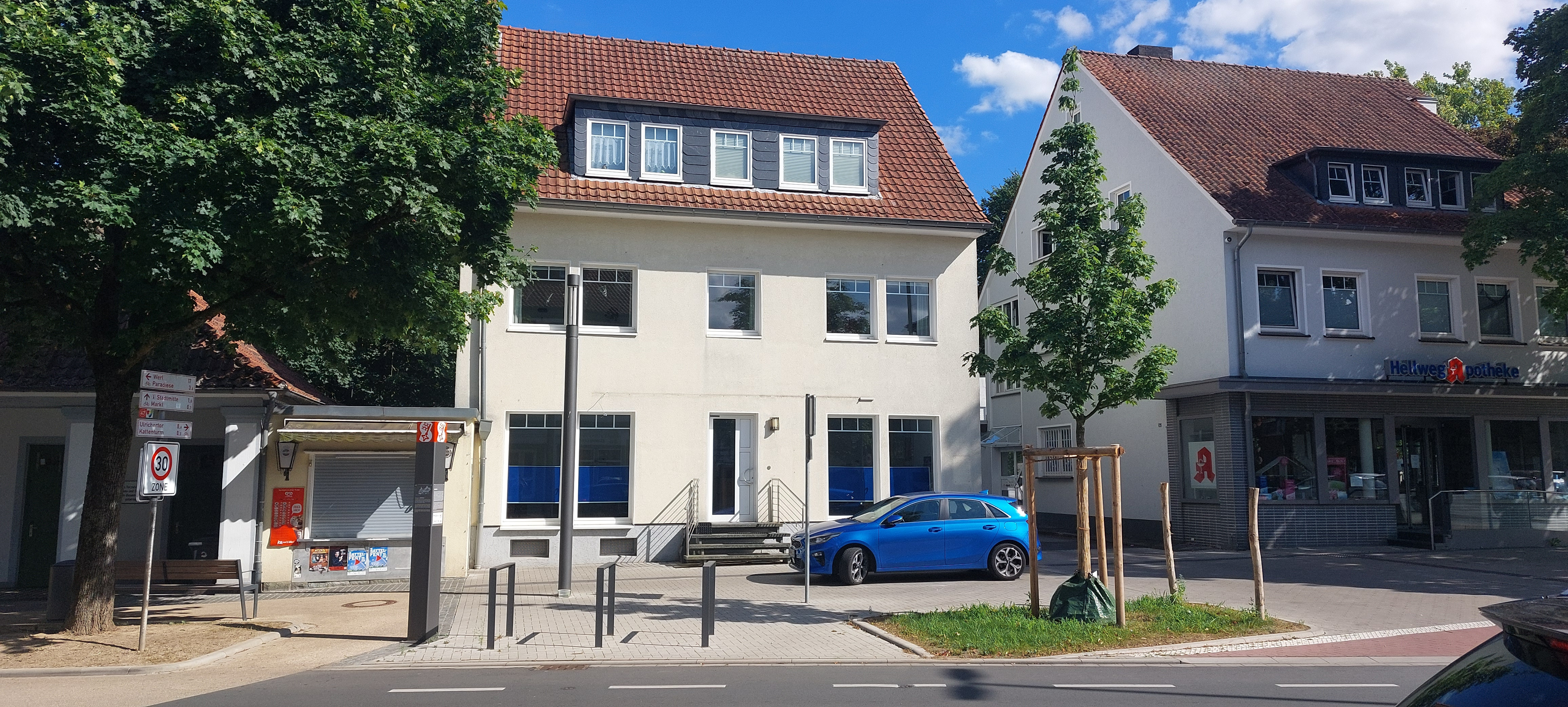 Helles zweistöckiges Gebäude mit Mansarddach neben einer Apotheke an einer Straße mit Parkplätzen und einem blauen Auto.