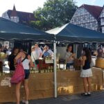 Zwei junge Frauen stehen vor einem Marktstand mit Honiggläsern auf einem gepflasterten Marktplatz, im Hintergrund historische Fachwerkhäuser und eine Baumreihe auf dem Soester Bauernmarkt.