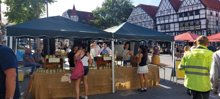 Zwei junge Frauen stehen vor einem Marktstand mit Honiggläsern auf einem gepflasterten Marktplatz, im Hintergrund historische Fachwerkhäuser und eine Baumreihe auf dem Soester Bauernmarkt.