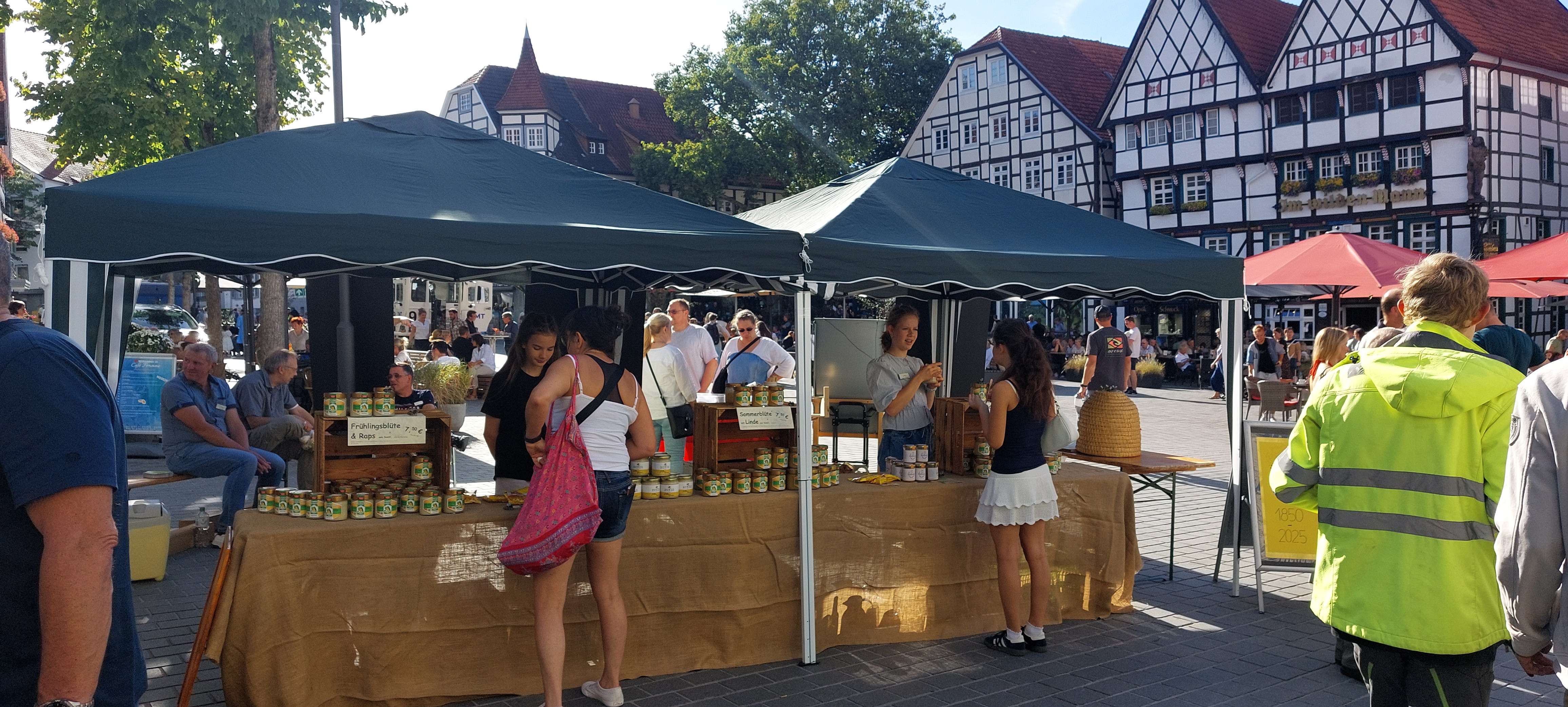 Zwei junge Frauen stehen vor einem Marktstand mit Honiggläsern auf einem gepflasterten Marktplatz, im Hintergrund historische Fachwerkhäuser und eine Baumreihe auf dem Soester Bauernmarkt.