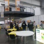 Einblick in einen gut besuchten Messestand auf der "Echter Deutscher Honig"-Ausstellung, mit Besuchern, die sich an Tischen unterhalten und Informationsstände betrachten, darunter ein Stand mit der Aufschrift "100 Jahre Echter Deutscher Honig".