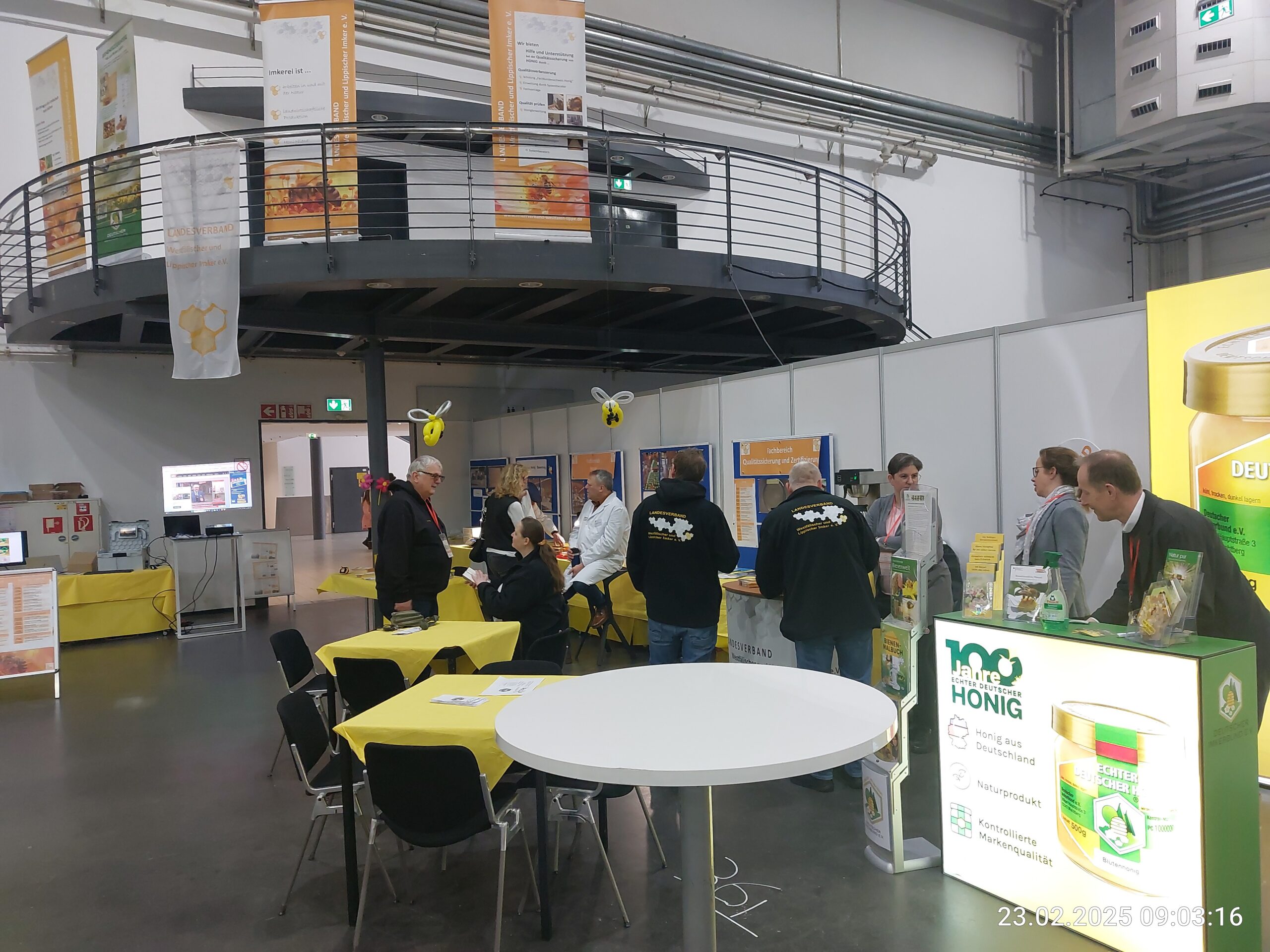 Einblick in einen gut besuchten Messestand auf der "Echter Deutscher Honig"-Ausstellung, mit Besuchern, die sich an Tischen unterhalten und Informationsstände betrachten, darunter ein Stand mit der Aufschrift "100 Jahre Echter Deutscher Honig".