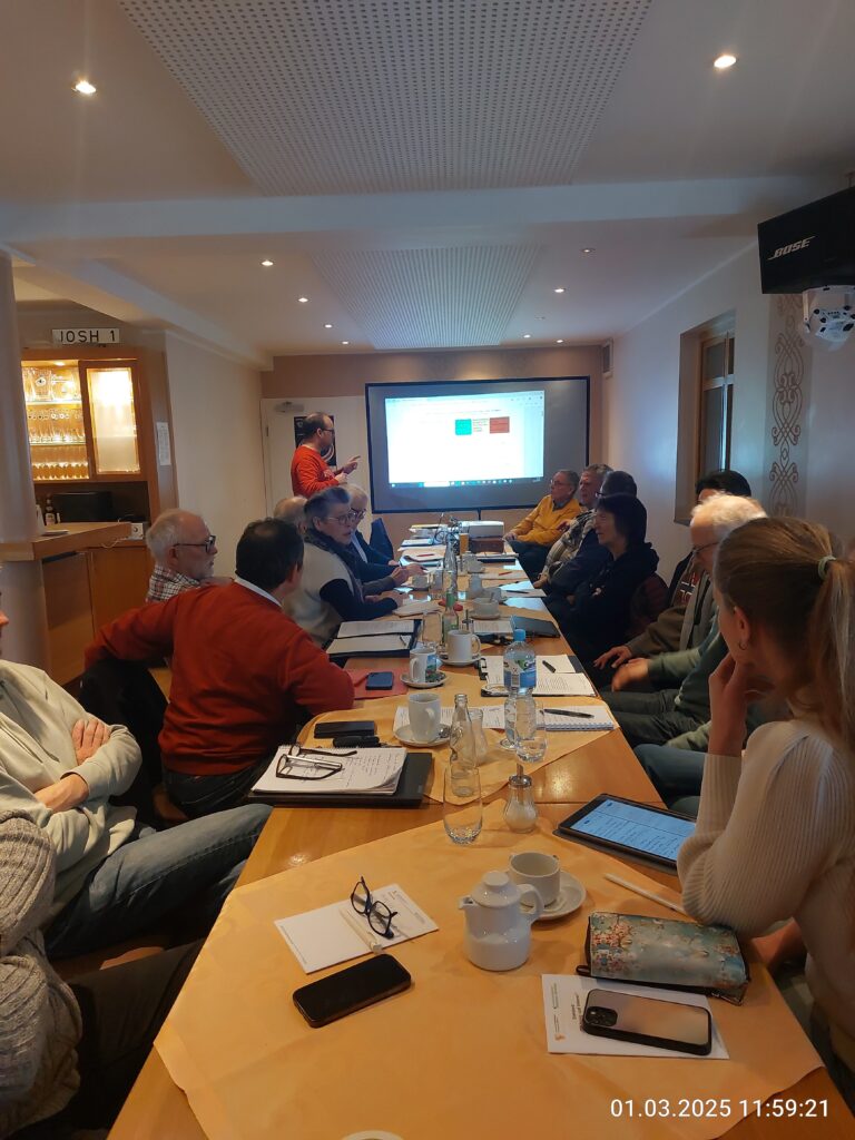 Foto eines Imker-Workshops des Landesverbandes Westfälischer und Lippischer Imker in einem gut beleuchteten Konferenzraum. Am vorderen Ende des langen Tisches, an dem etwa 14 Teilnehmer sitzen, steht ein Referent in einem orangeroten Hemd und zeigt mit einem grünen Laserpointer auf eine Projektionsleinwand mit Datenvisualisierungen. Auf dem Tisch sind klare Schilder mit der Aufschrift „IMKERBUND WESTFALEN-LIPPE“ und einem Programmhinweis „WORKSHOP IMKEREI 4.0: DIGITALISIERUNG IM BETRIEB - Westfälische Imker“ zu sehen. Auch Flaschen mit dem „D.I.B.“-Logo sind aufgestellt.