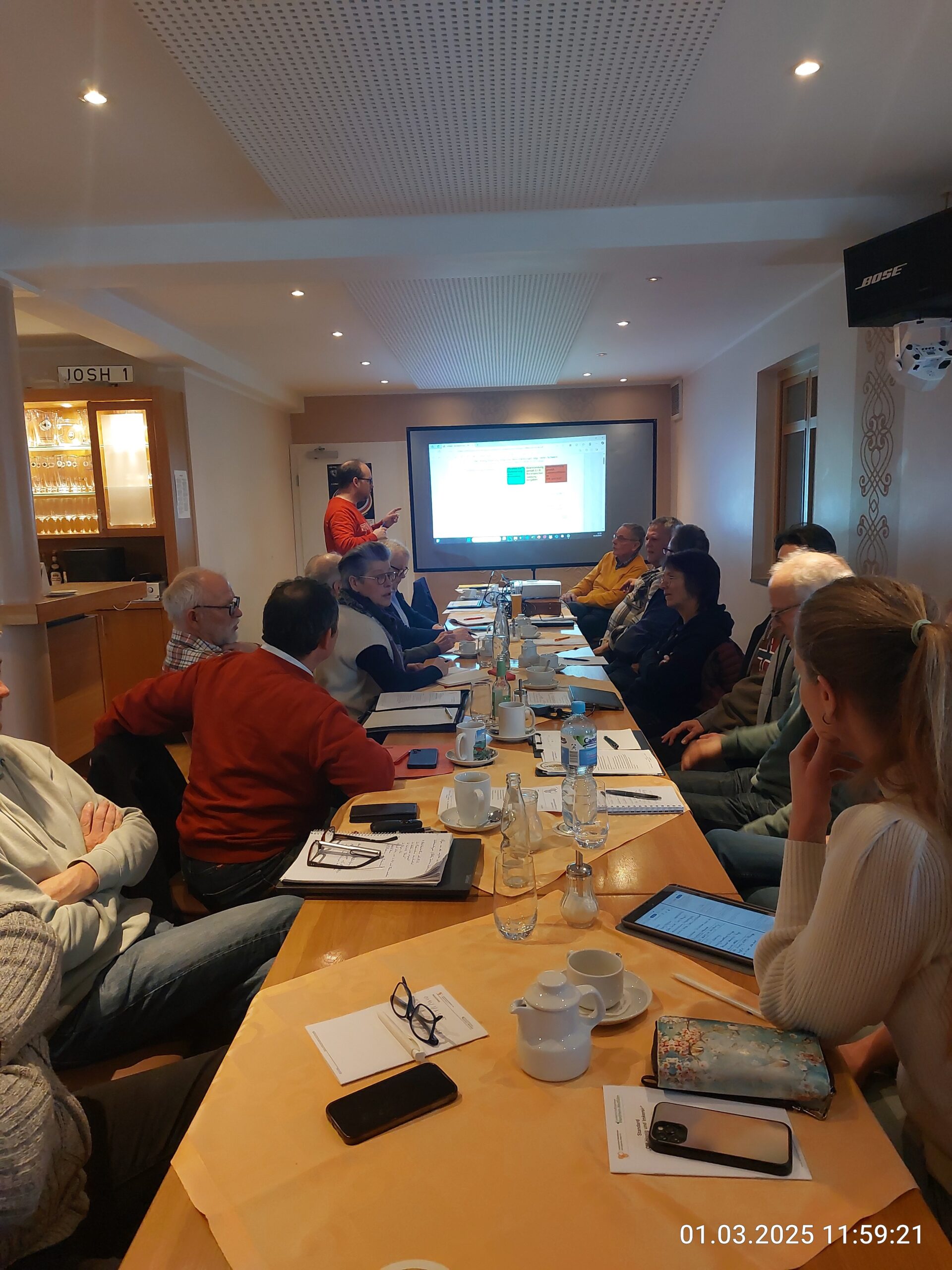 Foto eines Imker-Workshops des Landesverbandes Westfälischer und Lippischer Imker in einem gut beleuchteten Konferenzraum. Am vorderen Ende des langen Tisches, an dem etwa 14 Teilnehmer sitzen, steht ein Referent in einem orangeroten Hemd und zeigt mit einem grünen Laserpointer auf eine Projektionsleinwand mit Datenvisualisierungen. Auf dem Tisch sind klare Schilder mit der Aufschrift „IMKERBUND WESTFALEN-LIPPE“ und einem Programmhinweis „WORKSHOP IMKEREI 4.0: DIGITALISIERUNG IM BETRIEB - Westfälische Imker“ zu sehen. Auch Flaschen mit dem „D.I.B.“-Logo sind aufgestellt.