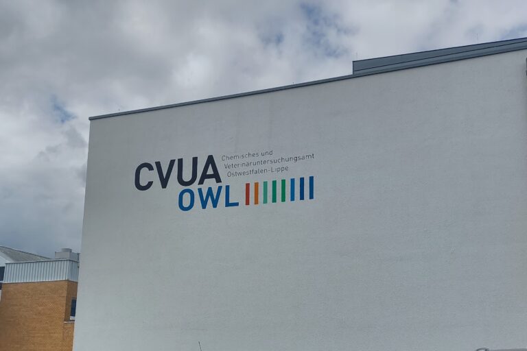 Außenansicht des Dienstgebäudes des CVUA OWL in Detmold mit Parkplatz, einem markanten türkisfarbenen Opel Corsa B mit Bielefelder Kennzeichen und dem CVUA-Logo an der Fassade.