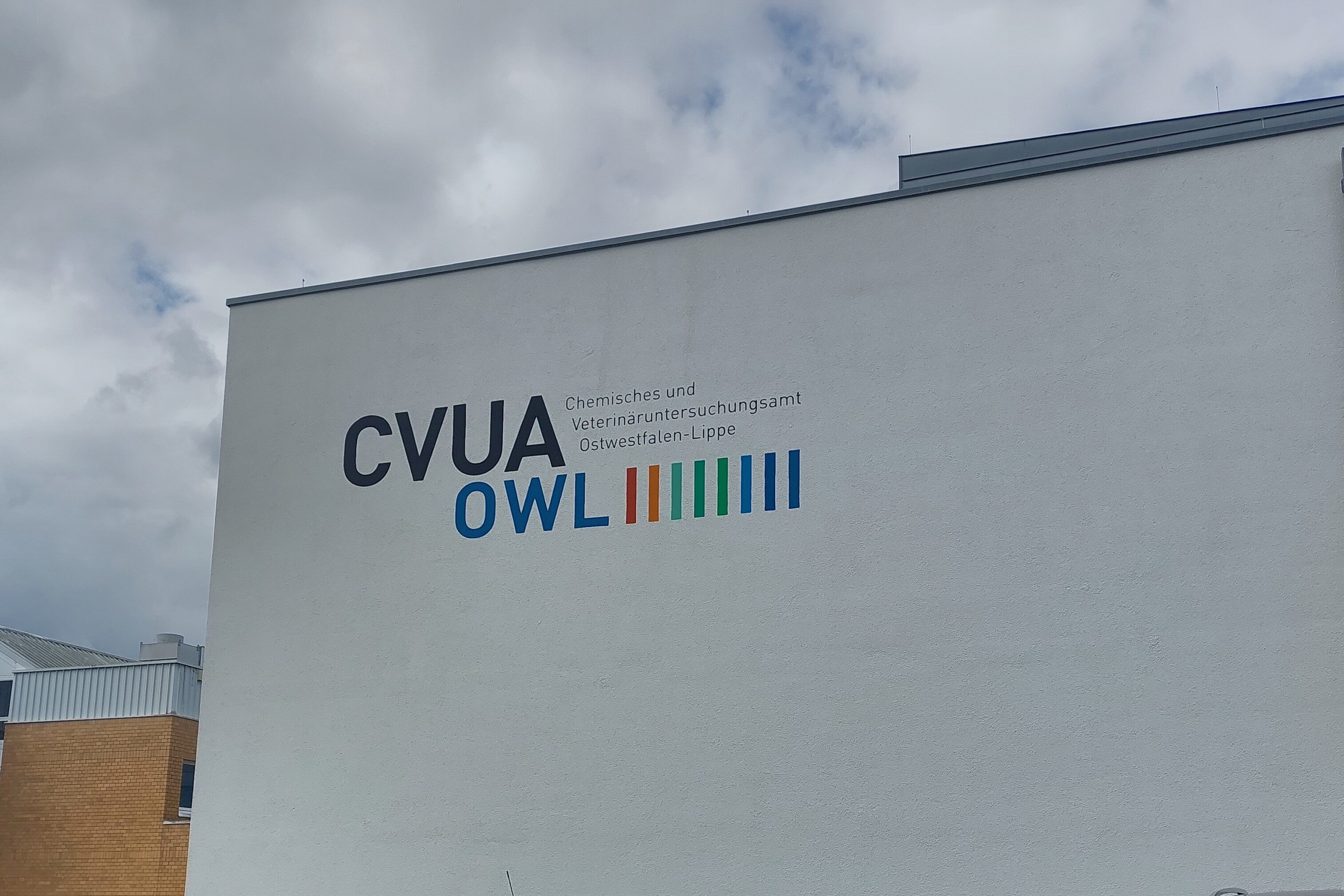 Außenansicht des Dienstgebäudes des CVUA OWL in Detmold mit Parkplatz, einem markanten türkisfarbenen Opel Corsa B mit Bielefelder Kennzeichen und dem CVUA-Logo an der Fassade.