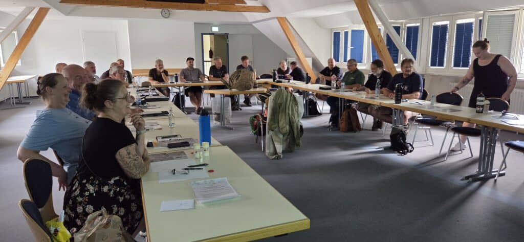 Teilnehmende des Seminars zur Vespa velutina Nestentfernung an einer U-förmigen Tischreihe mit einem ausgestellten Hornissennest auf Haus Düsse.