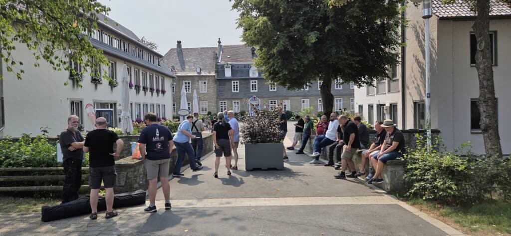 Teilnehmende des Vespa-velutina-Nestentfernerkurses stehen und sitzen bei Sonnenschein im Außenbereich und Innenhof von Haus Düsse.